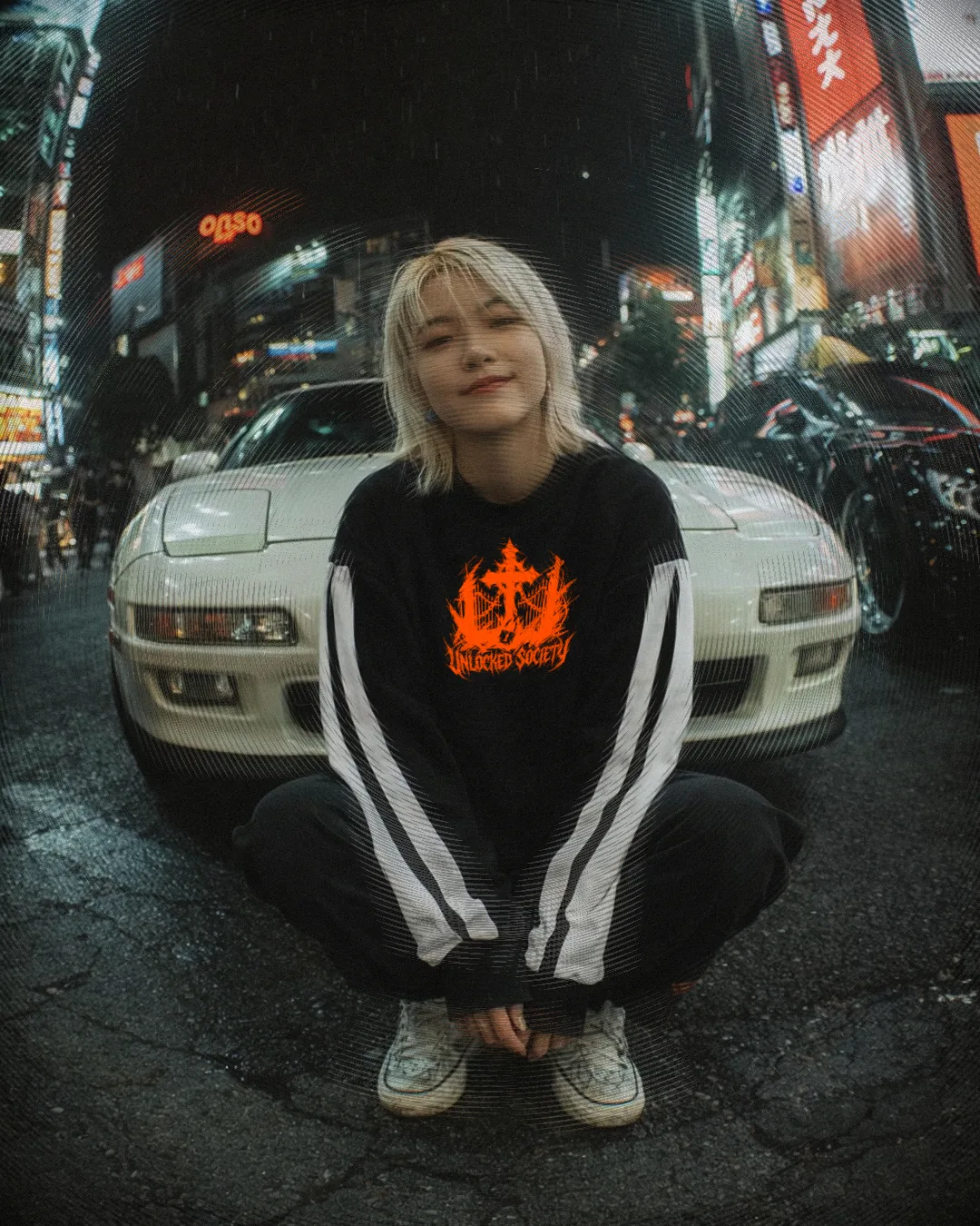 Campaign photograph shot on a fisheye lens: a person in a branded sweatshirt crouching in front of a white sports car on a neon-lit street at night.